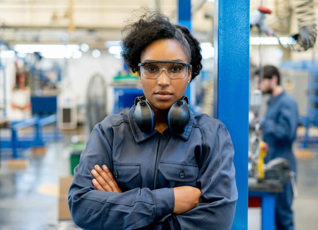 A young woman stands with arms crossed inside a manufacturing facility.