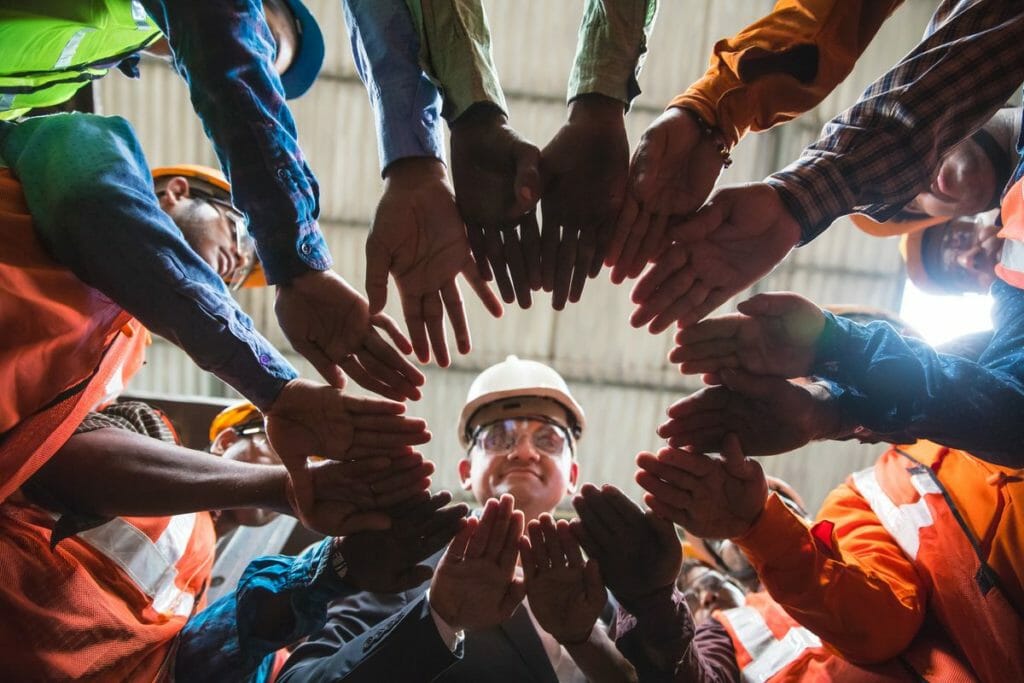 A team at a manufacturing plant circles up and puts their hands in the middle of the huddle.