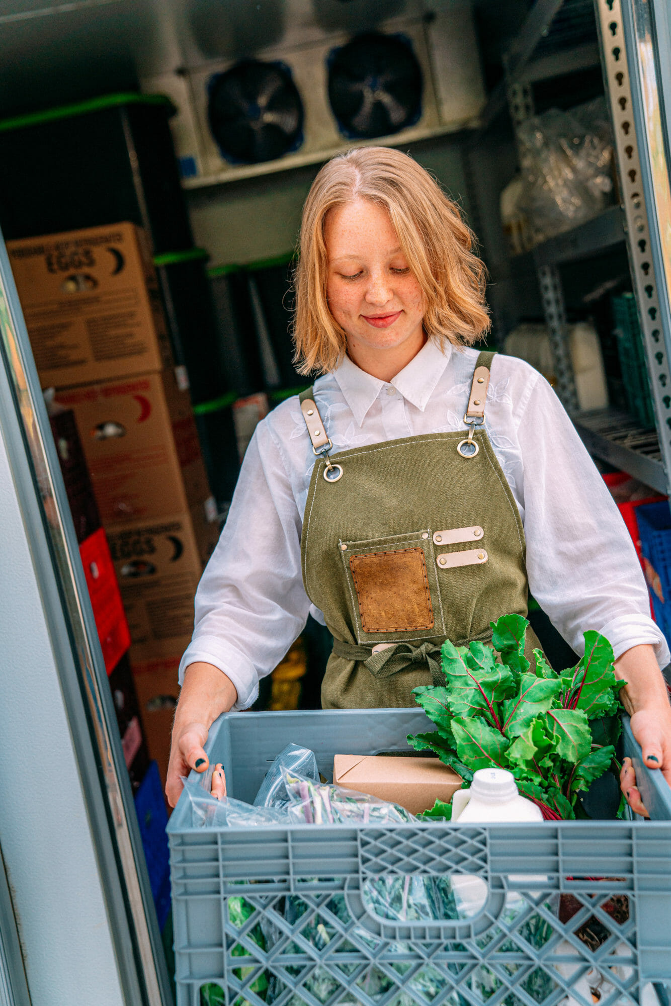 Portrait of a Happy Young Caucasian Woman Standing at the Door of a Walk-In Refrigerator Holding a Basket of Delicious, Healthy, Organic Produce at a Local Small Business Farm-to-Table Supplier in Colorado